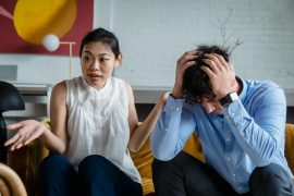 A couple sitting on a couch during a tense conversation, with a man holding his head while his partner gestures, illustrating emotional shutdown and communication challenges addressed in men’s counseling in Colorado Springs, CO.