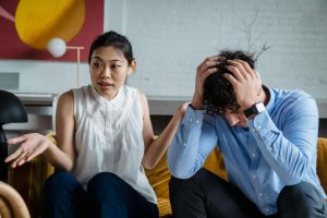 A couple sitting on a couch during a tense conversation, with a man holding his head while his partner gestures, illustrating emotional shutdown and communication challenges addressed in men’s counseling in Colorado Springs, CO.