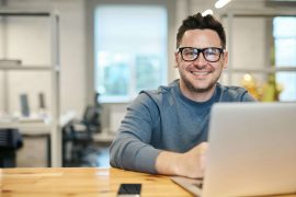 Man smiling while using a laptop in a bright workspace, showing the flexibility and ease of online anxiety counseling for men in Colorado Springs, CO.
