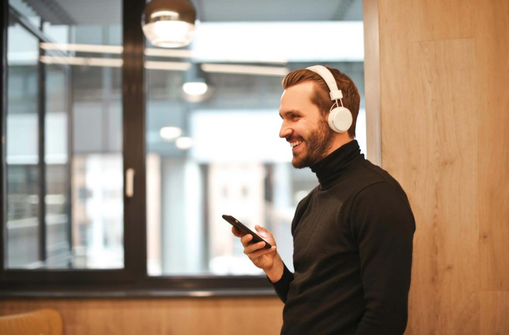 Man wearing headphones and smiling while using his phone indoors, representing support and connection with a therapist for men in Colorado Springs, CO.