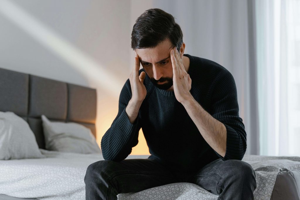 Man sitting on the edge of a bed with his hands at his temples, illustrating internalized grief and emotional overwhelm explored in grief counseling for men in Colorado Springs, CO.