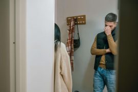 Man and woman standing in a hallway while the man covers part of his face with his hand and closes his eyes, representing emotional shutdown explored in stress management therapy for men in Colorado Springs, CO.