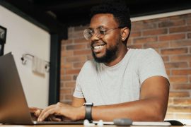 Man smiling while using a laptop, representing connection and support through online therapy for men in Colorado Springs, CO.